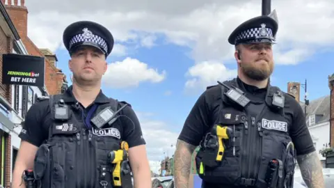 Alex Dunlop/BBC Two uniformed police officers walking down a high street. They are wearing black stab vests over black tops. There is a yellow Taser in each of their jackets and they have hats on their heads.