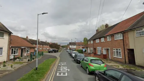 A Google street view image of Elgar Road, Hull. The street is narrow and has double yellow lines running down the left side. A row of parked cars are visible on the right side of the road. Rows of houses, front gardens, telegraph poles and street lights can also be seen on both sides of the image.