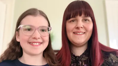 Family Handout A young girl (left) with brown hair and pink glasses smiles at the camera. She's wearing a navy tee shirt.
To her right her mother is also smiling. She has red hair. 