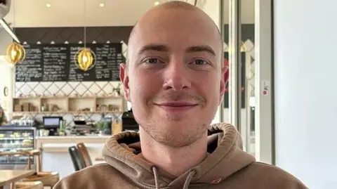 Eli Sweeting. He has close shaven hair and stubble on his chin. He is pictured wearing a brown hoodie and a nose ring. He is looking directly at the camera and smiling.