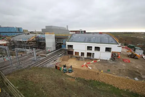 GTR Cambridge South railway station seen from the outside, perhaps a semi-ariel view. Train lines run into the main station undercover, while on the right a new white building is being constructed, with a curved perspex roof. There are a few safety barriers scattered around and a JCB in the distance. Behind the station are what look like offices/hotels and industrial buildings.