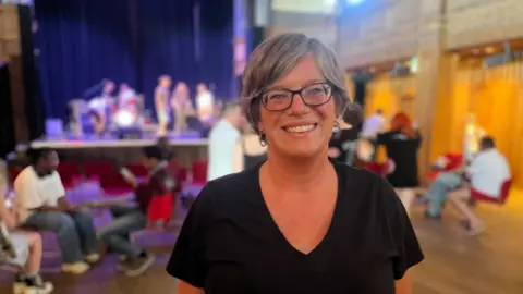 Qays Najm/BBC Jacqui MacKay is wearing a black V-neck top and is wearing dark rimmed spectacles. She has medium long blond highlighted hair, and she is smiling. She is standing inside St George's Theatre and there are people on stage and an audience on chairs.