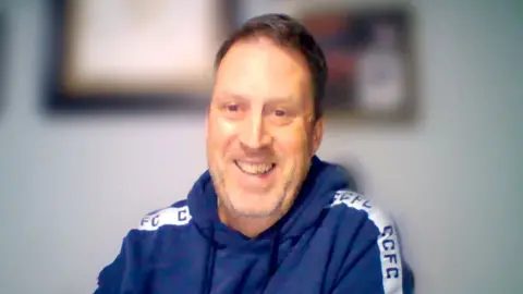 Mark Rushworth, a man with brown hair, is weating a blue hoodie which has white CCFC branding along the side on white stripes. There are paintings on a wall behind him, the background is blurred.