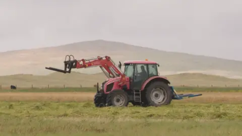 A red tractor crossing a green and brown field. The driver is a man wearing a hat. There's a hill in the background and clouds in the sky.