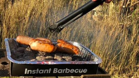 Getty Images Sausages seen cooking on a disposable barbecue with black tongs about to pick one up. A long grassy area is visible in the background