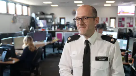 Ant Saddington/BBC A police officer in uniform. He is wearing glasses and standing in a police control room, with desks of computers behind him. 