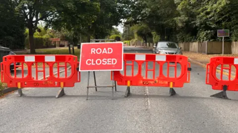 BBC A large, plastic, red barrier blocks a road with a "Road Closed" sign in the middle. Beyond this is an empty road with one single, silver-coloured car and trees and grass beside the road