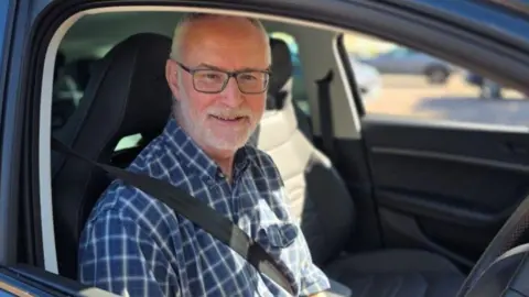 Fran Whymark, a man in a blue chequered shirt who is sitting inside a car in the driving seat and is wearing a seat belt. 
