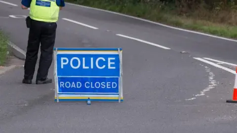 Getty A blue road sign that says 'police, road closed'. It is on a road with an officer stood near, you cannot see their face.