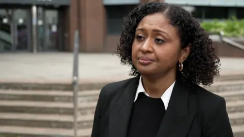 A woman with short dark curly hair and wearing a suit jacket with white collar protruding stands in front of the court.