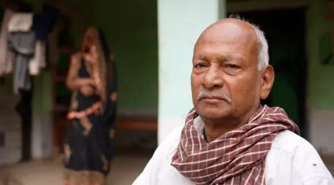 A man wearing a white shirt ad red checkered scarf looks into the camera
