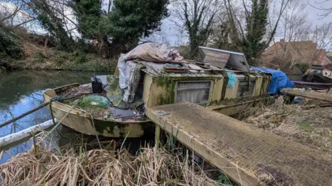 A badly run down narrowboat, which is partially covered in algae or moss is listing slightly at the side of a canal.