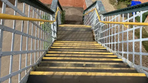 Vikki Irwin/BBC A shot down bridge steps looking towards the ground. The steps are black and the edges are yellow. They are lined by a grey handrail.