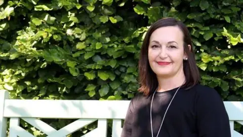 A smiling woman in a black top sat on a bench in front of a hedge