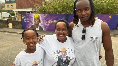 Irene stands between her son Elliot and a younger child. She and the younger child are wearing white t-shirts with a picture of Eddie on them and his name. The three are holding each other and smiling brightly in front of the mural. 