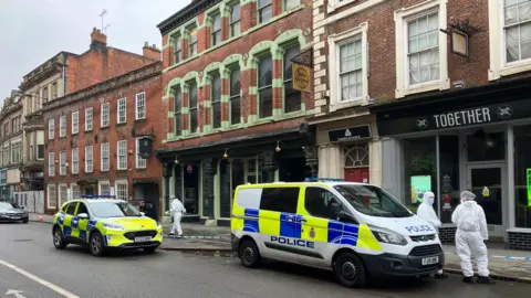 A street in the old quarter of Derby, with two police vehicles and people dressed in white forensic suits