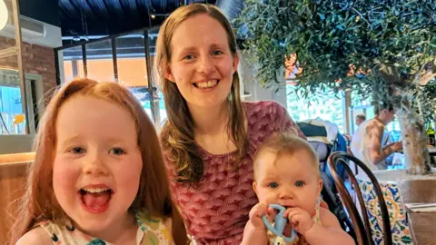 Maria Bennett An image of Maria Bennett with her two children. One child is a baby with a teething toy in its mouth and the eldest child is around 4-years-old and has red hair. Maria and her older daughter are both smiling at the camera. They look like they are in a restaurant setting. 