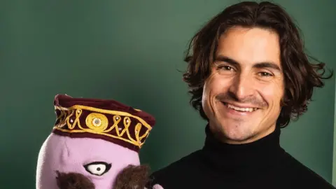 cruzstudios Kieran R. Leonard wearing a black turtleneck with short dark curly hair smiling at the camera with a puppet in his hand but it is slightly out of shot of the camera.