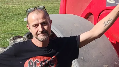 David Evans standing next to an agricultural vehicle in a field. He has short hair and a goatee beard and is wearing a red and black AC DC t-shirt 