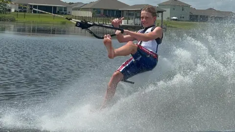 Matilda Mulcahy Olly Moore is pictured in a white and blue suit holding onto a rope on the back of a boat and he skis using just his feet. He has one foot up in the air as water sprays behind him.