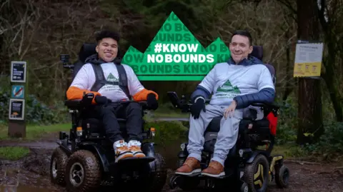 GFX Media Maxwell McKnight and Josh Wintersgill side by side in their wheelchairs in a woodland setting with a sign carrying the name of their challenge, Know No Bounds