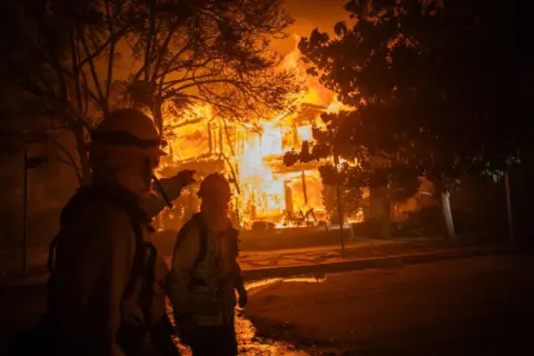 Getty Images Firefighters walk by house on flames in the in the Pacific Palisades neighbourhood of Los Angeles