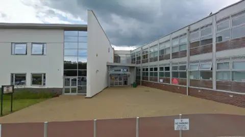 A Google Streetview image of a school on a day with grey skies. It is a rendered building with glass windows along the top and bottom levels. 