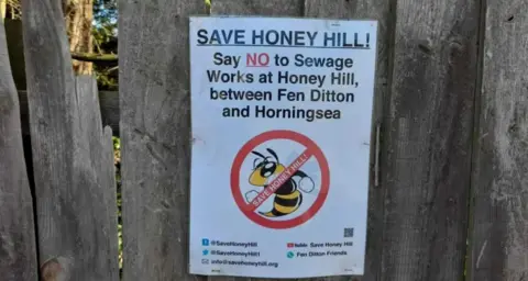 Local Democracy Reporting Service A poster on the wooden fence. The poster reads "Save Honey Hill! Say NO to sewage works at Honey Hill between Fen Ditton and Horningsea". The text is accompanied by an image of a yellow and black bee, with a red circle and line through the image.  