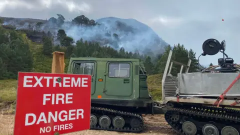 Ronan Dugan An outdoors vehicle parked in front of an area of wildfire in the Cairngorms. There is white smoke rising from behind a tree line in front of a hill in the shadows. In the foreground is a red sign with the words "extreme fire danger no fires" in white writing.