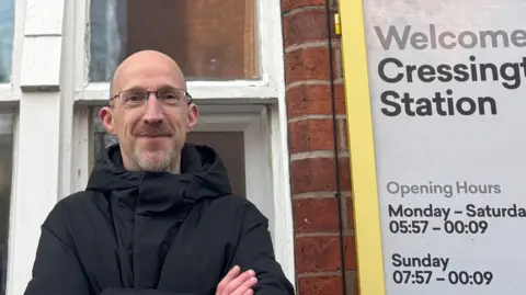 Councillor Richard Clein with a grey beard wearing glasses and a black coat stands at Cressington railway station with a yellow sign showing opening hours to his right.