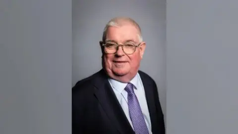 John Tizard with white hair and round glasses. He has a black suit on, with a light blue shirt and a purple spotted tie. It is an official photograph and he is standing in front of a grey background.