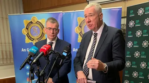 PA Media Two men stand in front of several microphones. Behind the man on the left, who is shorter and wearing glasses, there is An Garda Síochána branding. Behind the man on the right, there is PSNI branding. 