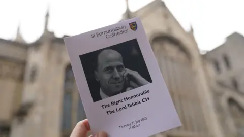 PA Media An order of service is held up in front of St Edmundsbury Cathedral, it has a black and white image of Lord Tebbit and details of the date and timings of the funeral service.