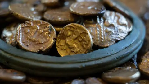 A close-up of a pile of 933 Iron Age gold coins. It shows the rim of a vessel, surrounded by coins with more gold coins heaped within.
