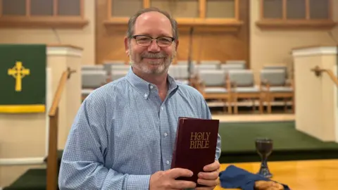 Pastor Tim Bostick smiling at the camera, he has dark brown hair and is wearing glasses. He is wearing a checked shirt and is holding a bible.