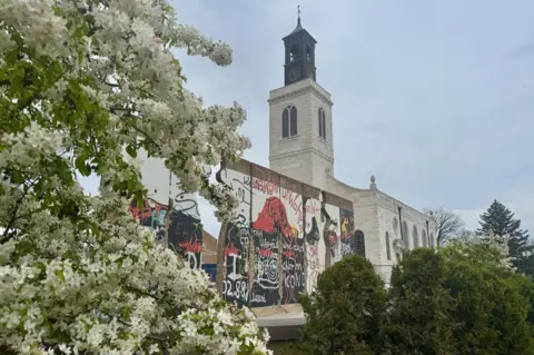 Tim Riley St Mary Aldermanbury as it is seen today alongside a sculpture featuring painted sections of the Berlin Wall with a flowering plant in the foreground