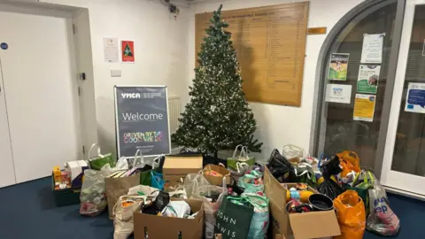YMCA Cheltenham Facebook A photo showing a collection of boxes and bags filled with gifts. They have been placed in front of a decorated Christmas tree, which is next to a welcome sign