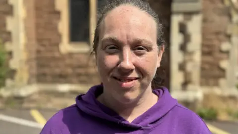 A woman with tied-back dark hair and a purple hooded top standing in the street