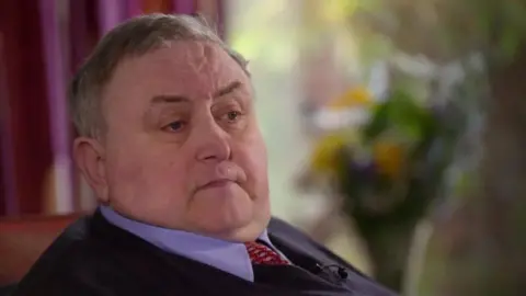 BBC Alan McQuillan, a man wearing a blue sweater over a blue shirt and red tie, sitting down