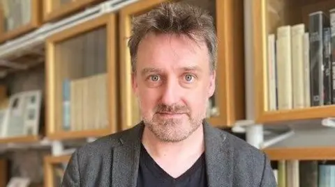 Petr Mackay is looking at the camera with shelves full f books behind him. He has short, grey/brown hair and is wearing a black T-shirt and a grey suit jacket.