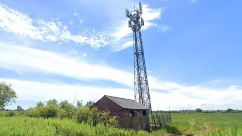A telecommunications mast stands in a field next to a small fenced off building.