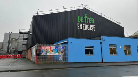 BBC The outside of a leisure centre. A red brick and black and blue corrugated iron building with a sign on the side reading Better Energise.