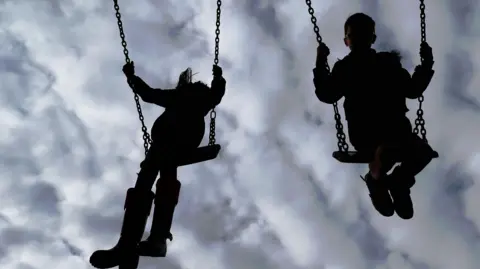 The outline of two children play on two swings. The swings are in mid-air and in the background is a cloudy sky. 