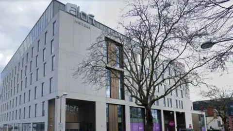 Google Exterior photo of Unite Hotel and Conference Center in Birmingham. It's a gray building with a lobby with a sign above the door bearing the name of the Aloft hotel chain that is based there.