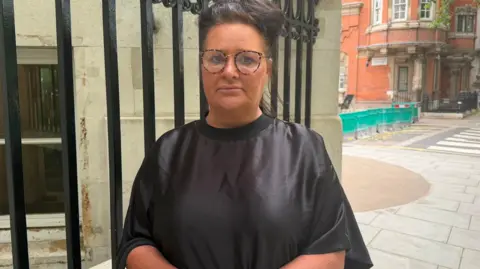 A woman with dark hair and glasses and wearing a smart black dress stands formally in the street. Her handbag is on her right arm.