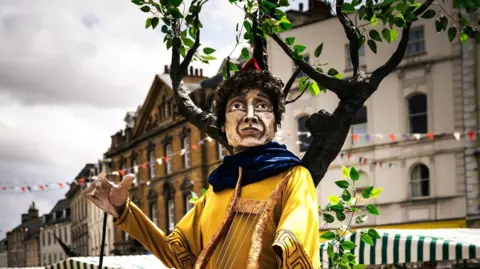 Alex Tabrizi The Orpheous puppet looks towards the camera. There is papier mache foliage behind him, and he is pictured on an old city street in Cirencester lined with bunting and market stalls. 