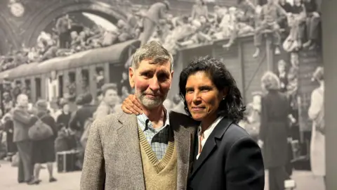 Clark McColme, who has a grey hair and beard and it wearing a mustard jumper and a tweed jacket, and his wife Beverly, who has dark hair and is wearing a dark blue formal jacket, standing in front of a black and white image in the exhibition.