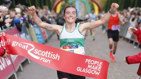 Great Scottish Run Biegarz w małym hałdzie, który czyta „Katie” oraz białą i zieloną kamizelkę z napisem „gali Harriers”, przecina linię wielkiego szkockiego biegu z podniesionymi ramionami. Taśma końcowa brzmi 