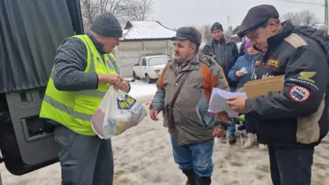 Gary Fear A man wearing a woolly hat, a long-sleeved top, grey trousers and a hi-vis vest hands out a bag of supplies to a waiting man.