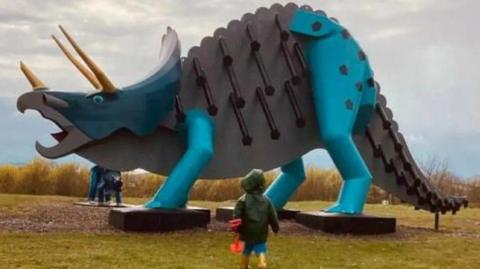 A small boy holding a spade approaches a giant steel dinosaur